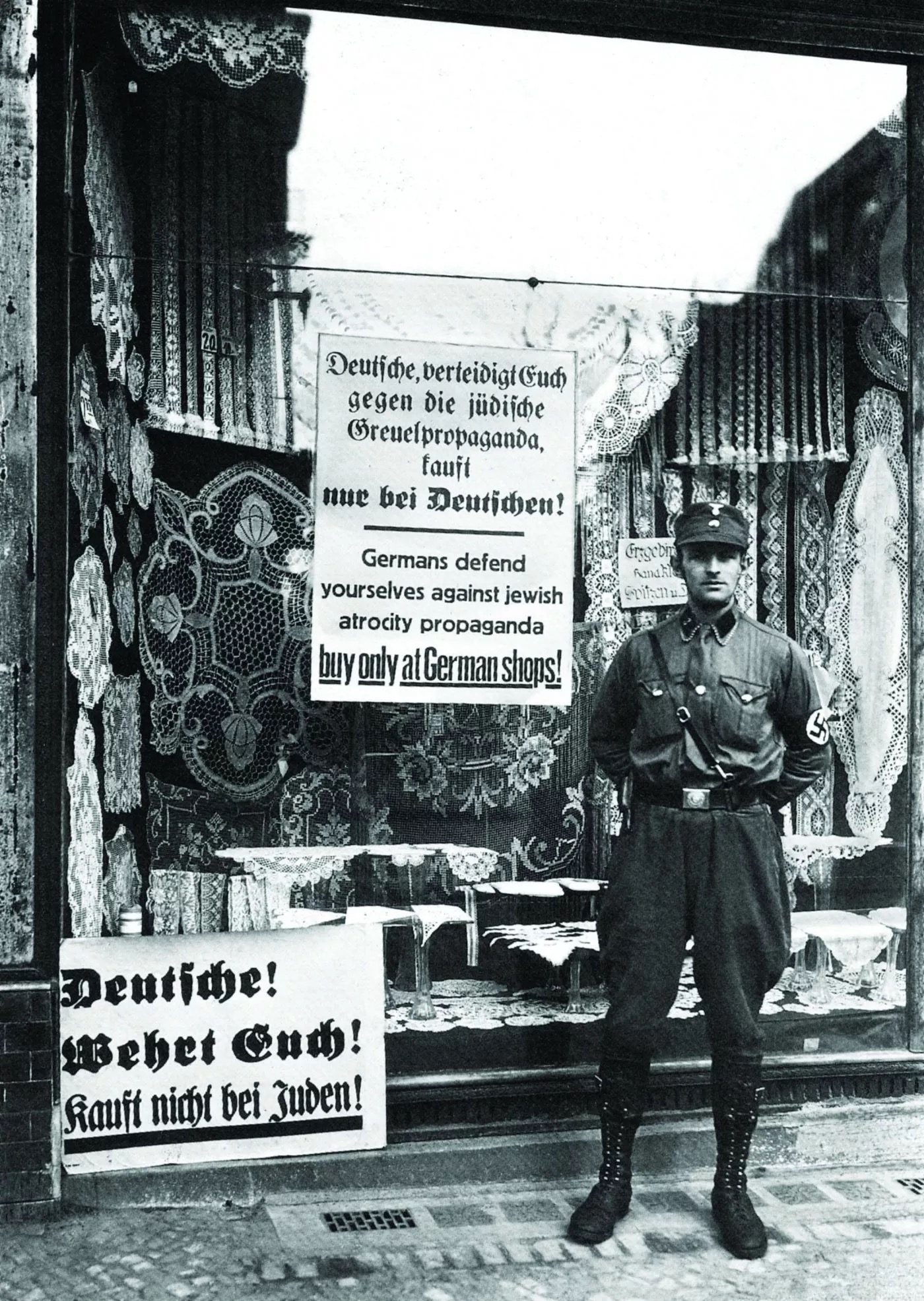 An SA (Sturmabteilung) brownshirt stands outside a Jewish-owned lace and crochet shop in Berlin during the Nazi boycott of 1 April 1933. Signs in the shop window read &ldquo;Deutsche! Wehrt Euch! Kauft nicht bei Juden!&rdquo; (&ldquo;Germans! Defend yourselves! Don&rsquo;t buy from Jews!&rdquo;).