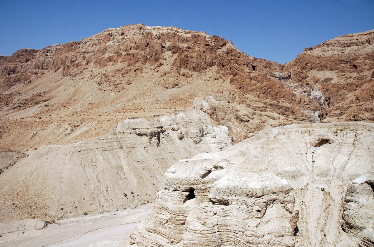 A wide view of the arid Judean Desert escarpment at Qumran, with eroded limestone cliffs in the foreground and the Dead Sea visible in the distance.
