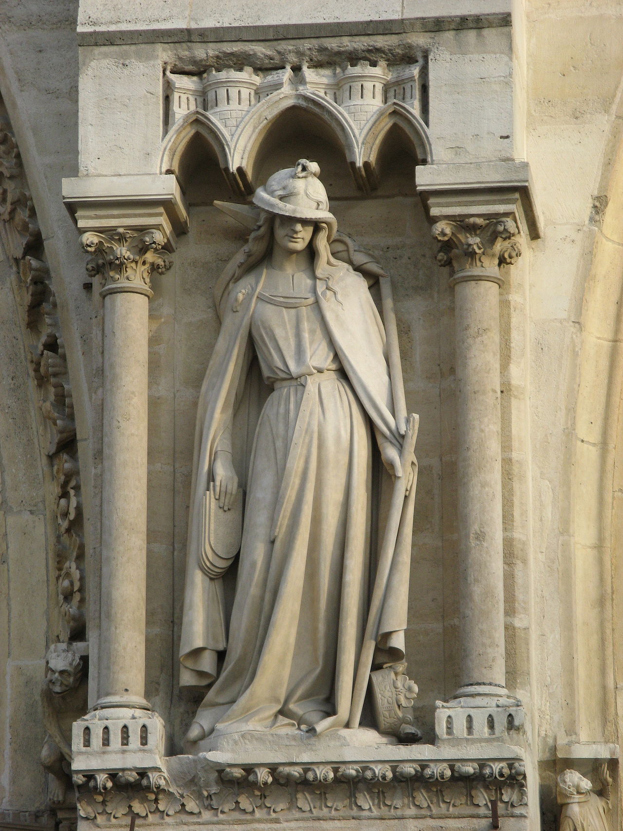 A medieval stone statue of a woman, elegantly carved, standing with a serpent coiled across her eyes, holding a broken staff, with tablets slipping from her lowered hand.