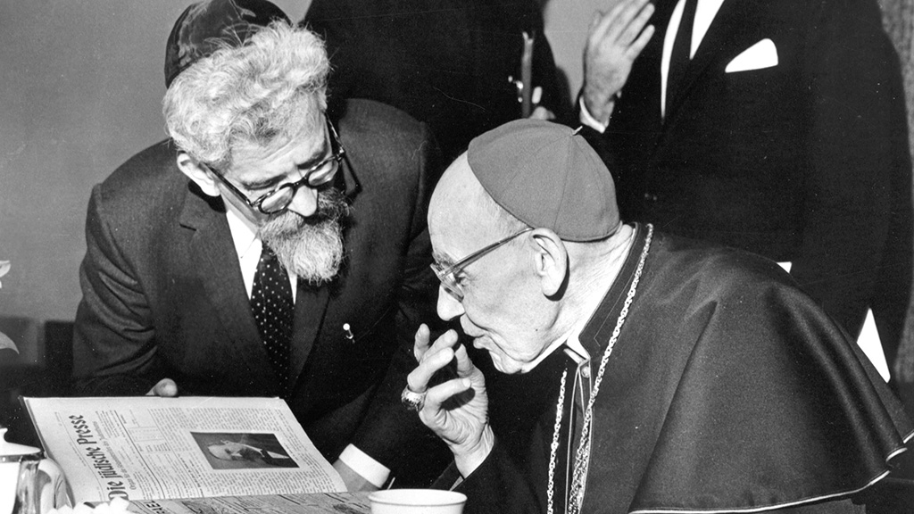 A black-and-white press photograph of two elderly men leaning close together over an open Jewish newspaper. On the left, a bearded man in a yarmulke and dark-rimmed glasses; on the right, a Catholic cardinal in a skullcap and robes with a pectoral cross, one hand raised mid-gesture.