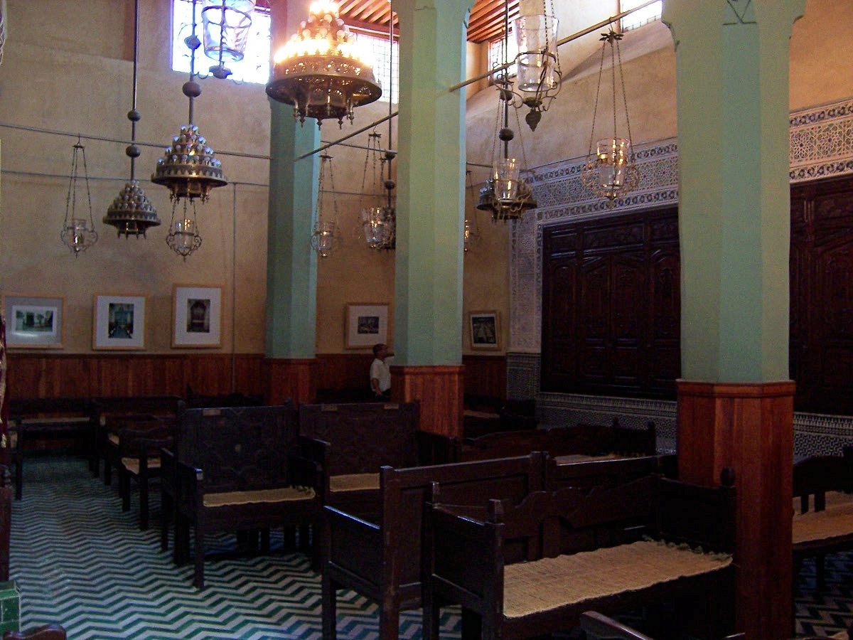 The interior of a small synagogue: green-and-white tiled floor, dark wooden benches, brass lamps hanging from a painted ceiling.