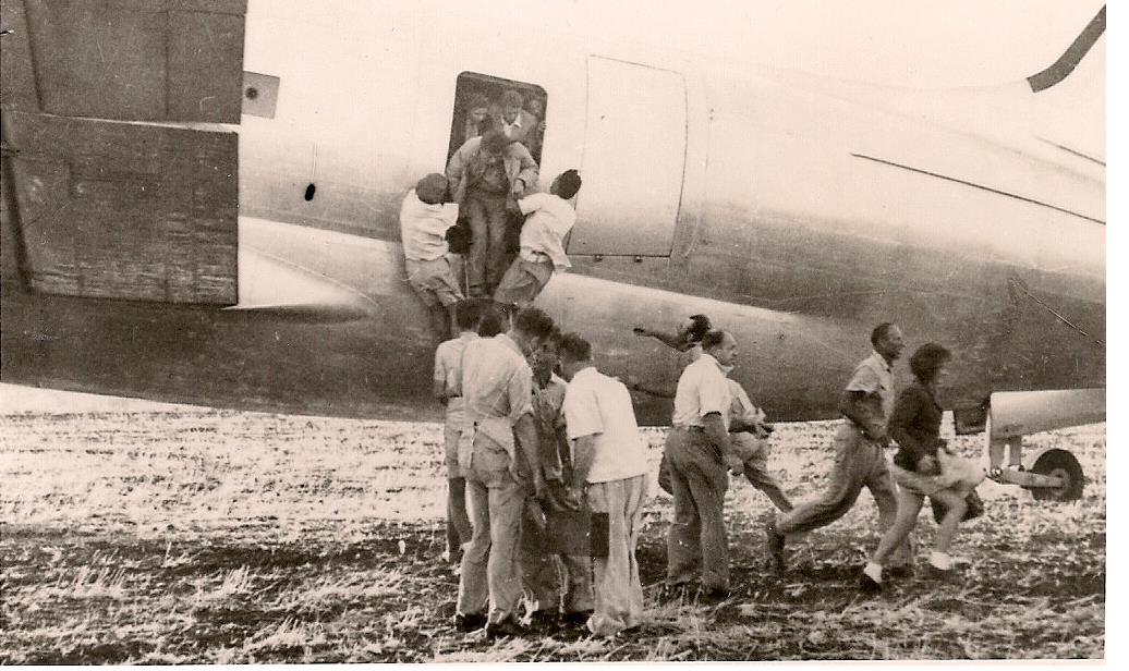 A black-and-white photograph of people climbing into the door of a propeller aircraft on a dirt airfield, carrying bundles, during the 1951 airlift of Iraqi Jews.