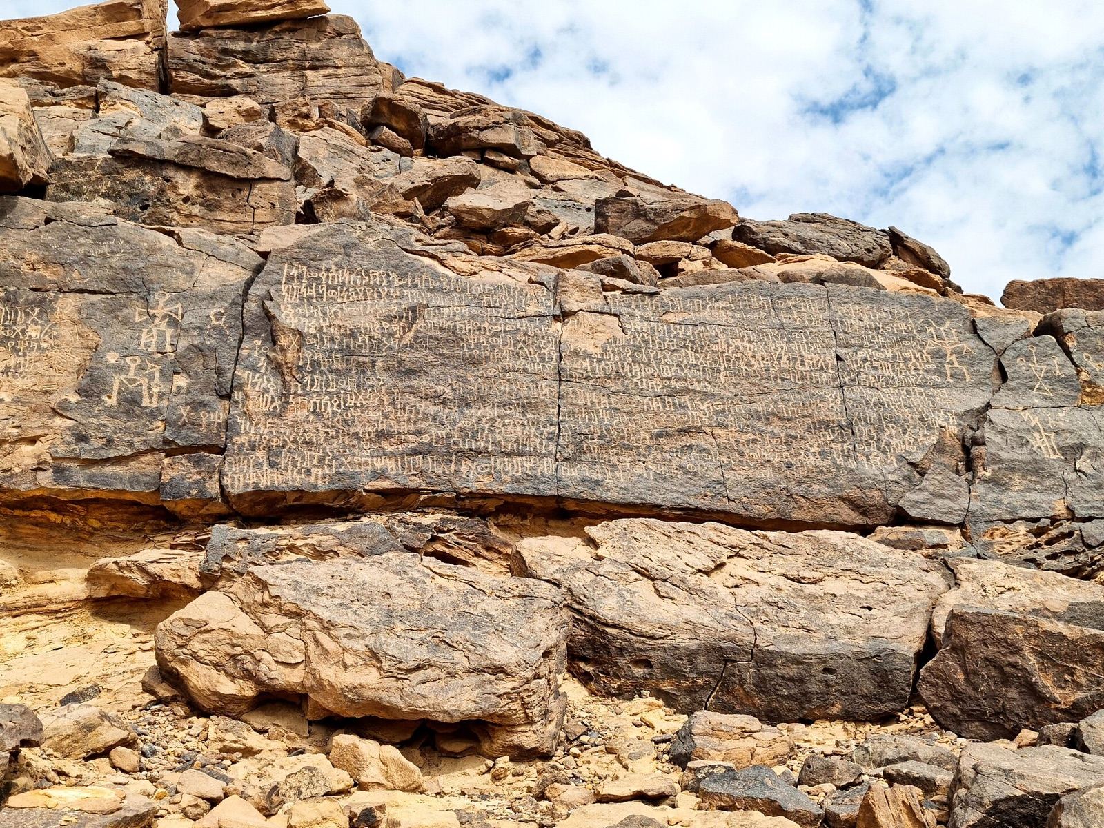A long ancient inscription carved in neat rows across a dark rock face in the desert, with small human figures carved beside it.