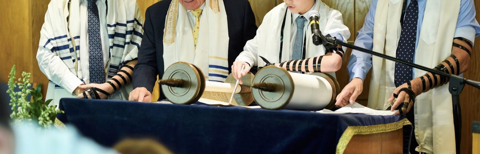 A bar mitzvah reads from an open Torah scroll with a pointer, flanked by men in prayer shawls