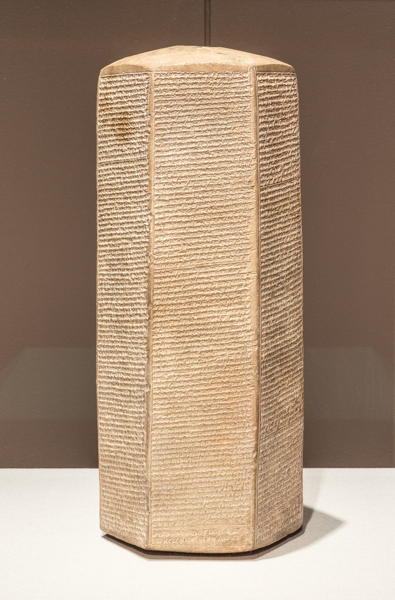 A six-sided baked-clay prism standing upright on a museum mount, every face packed top to bottom with rows of tiny wedge-shaped cuneiform writing.