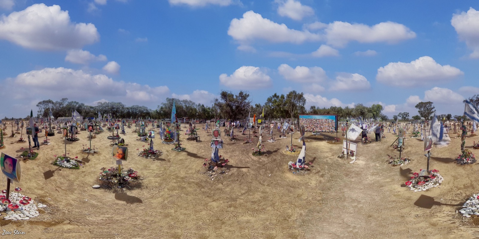 A wide view of the Nova festival memorial site near Re'im: photographs of the murdered mounted on poles among planted flowers, stretching across the open field where the festival stood.