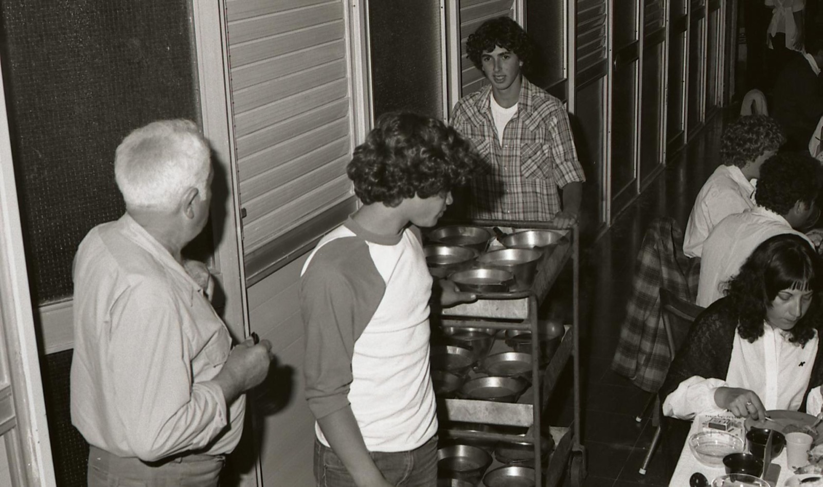 Teenagers serving a communal meal from a food cart in a kibbutz dining hall