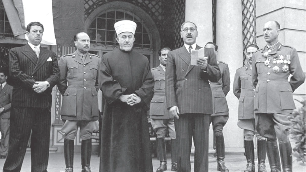 A black-and-white photograph of a row of men standing outdoors in front of an arched building. At center is a man in a white turban and dark robe with his hands clasped; beside him a man in glasses and a dark double-breasted suit reads from a sheet of paper held up in one hand. They are flanked by several men in military uniforms, one at the right wearing rows of medals.